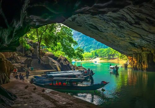 3. Boats docked inside a cave with an opening leading to a lush, green riverbank, where trees and rocky stairs create a scenic view.